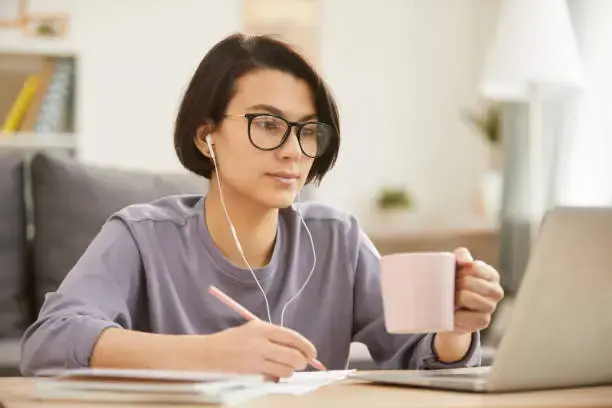 Lady holding a mug while and notetaking while looking at her laptop.
