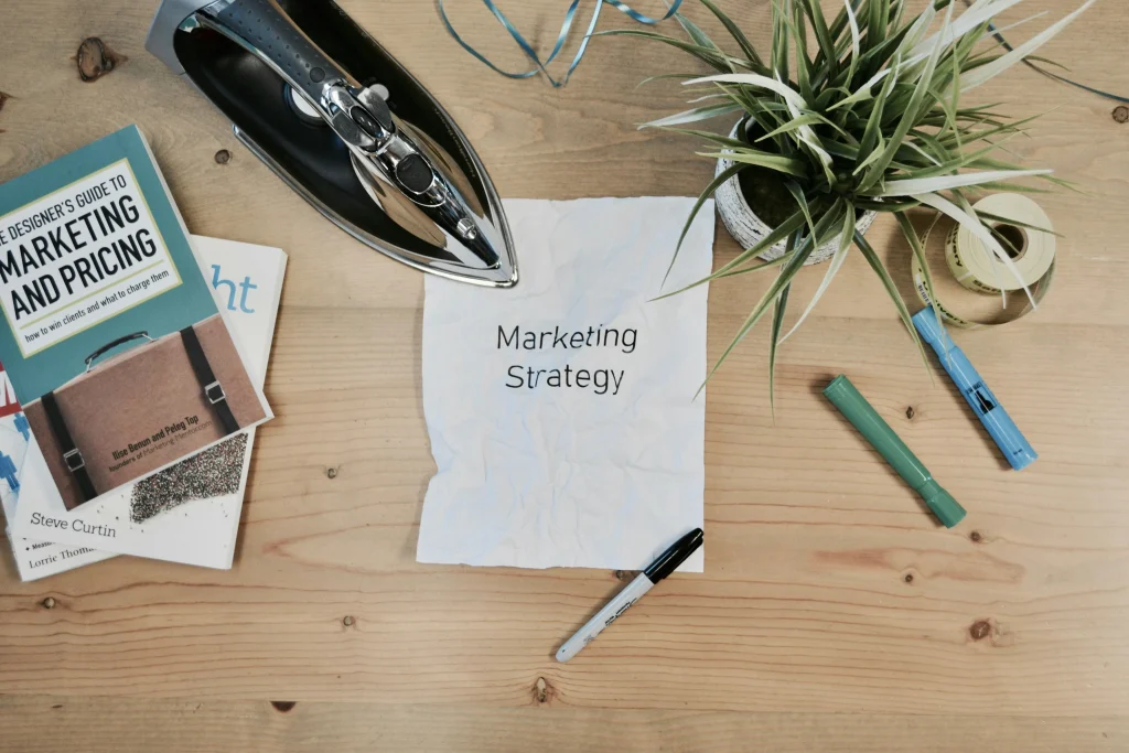 A crumpled paper with "Marketing Strategy" written on it lies on a wooden desk surrounded by a plant, markers, books on marketing, tape, and an iron.