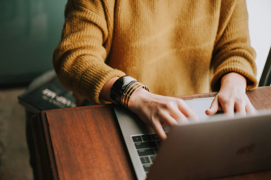Person typing on a laptop, wearing a yellow sweater and bracelets.