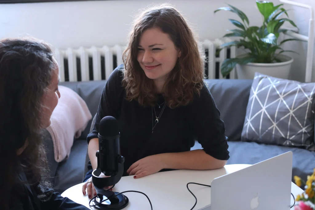 Two women collaborating with a microphone and laptop, suggesting a virtual or podcast setting.