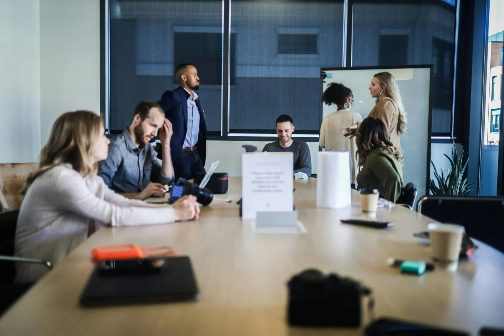 A group of people gathered in a conference room, some working at a table while others stand and talk, suggesting a casual team meeting or brainstorming session.