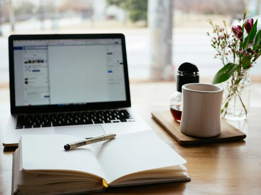 An open laptop sits on a desk beside a notebook with a pen, a coffee mug, and a small vase with flowers, creating a cozy workspace.